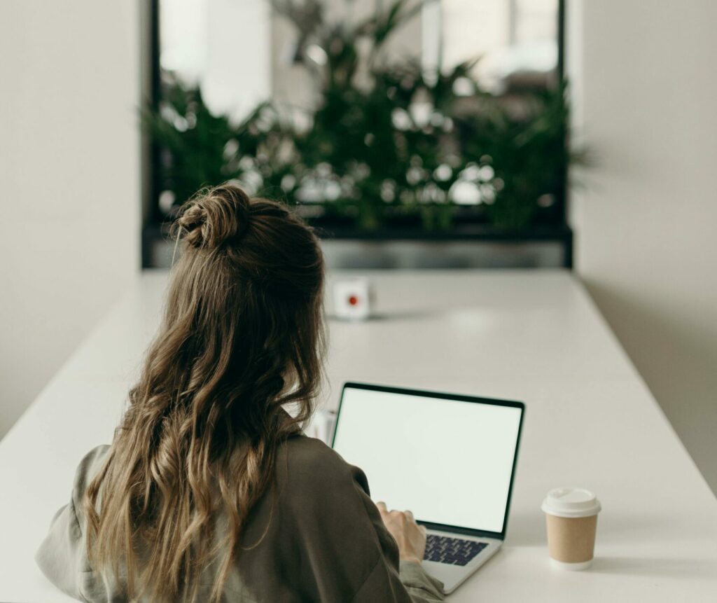 pexels-photo-4065624-4065624 A woman with curly hair works on a laptop, facing a window with plants, symbolizing remote work.