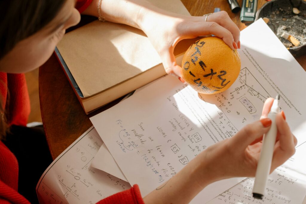 pexels-photo-4778676-4778676 Student studying math creatively using an orange and notes in warm indoor light.